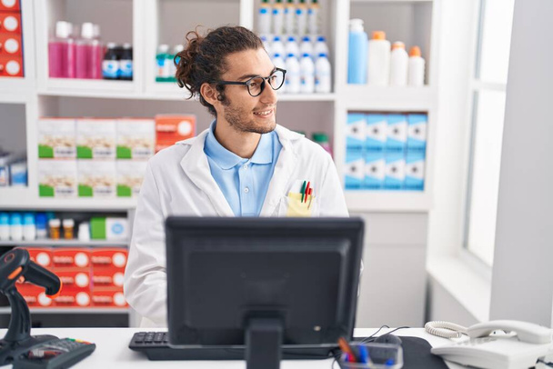 Молодий іспаномовний чоловік фармацевт посміхається впевнено використовуючи комп'ютер в аптеці
 - Фото, зображення