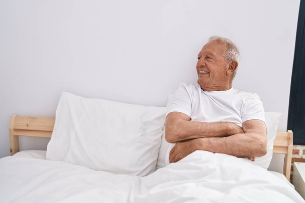 Ο ηλικιωμένος γκριζομάλλης χαμογελάει με αυτοπεποίθηση καθισμένος στο κρεβάτι στην κρεβατοκάμαρα - Φωτογραφία, εικόνα