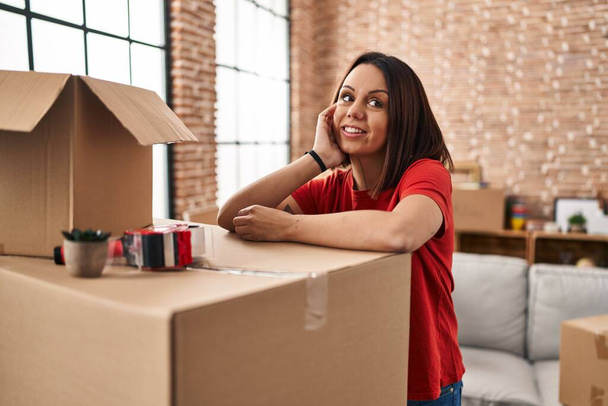 Junge schöne hispanische Frau lächelt selbstbewusst an Paket im neuen Zuhause angelehnt - Foto, Bild