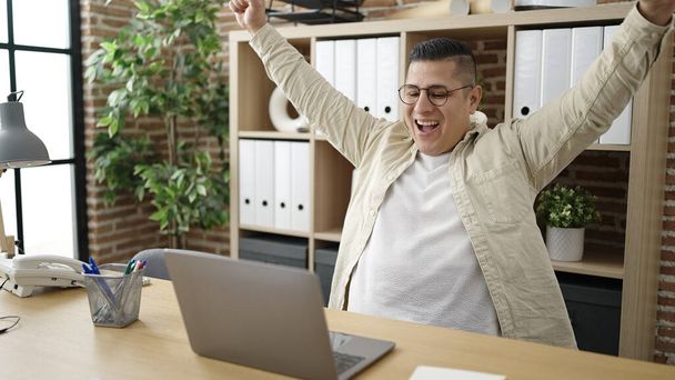 Jonge Spaanse man zakenman met behulp van laptop vieren op kantoor - Foto, afbeelding