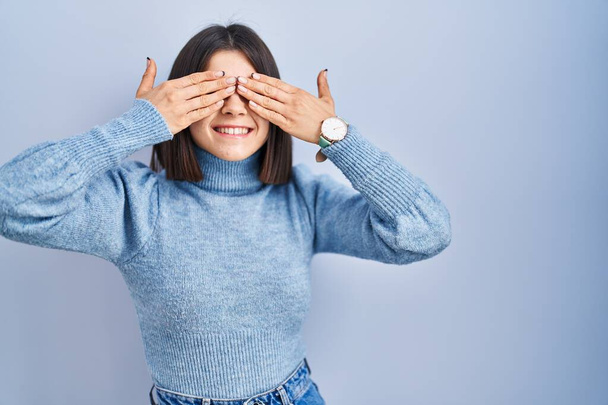 Jonge Spaanse vrouw die over een blauwe achtergrond staat en ogen bedekt met lachende, vrolijke en grappige handen. blind concept.  - Foto, afbeelding