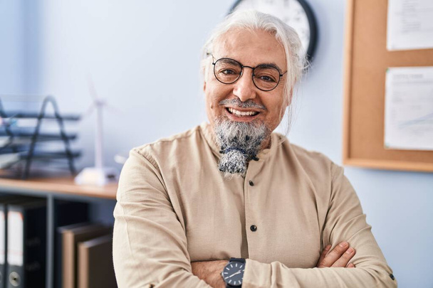 Hombre de pelo gris de mediana edad trabajador de negocios sonriendo confiado de pie con los brazos cruzados gesto en la oficina - Foto, Imagen
