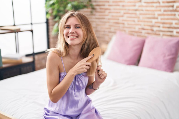 Young blonde woman combing hair sitting on bed at bedroom - Photo, Image