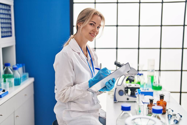 Nuori blondi nainen tiedemies kirjoittaa asiakirjaan laboratoriossa - Valokuva, kuva