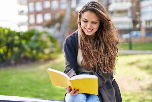 Jonge mooie Spaanse vrouw die boek leest zittend op bank in park - Foto, afbeelding