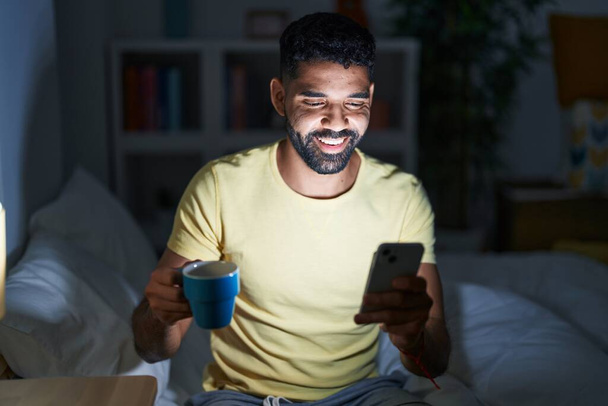 Jonge Arabier man met behulp van smartphone koffie drinken in de slaapkamer - Foto, afbeelding