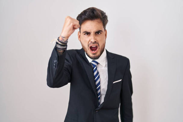 Junger hispanischer Mann mit Tätowierungen in Businessanzug und Krawatte, wütend und wütend die Faust reckend frustriert und wütend, während er vor Wut schreit. Wut und aggressives Konzept.  - Foto, Bild