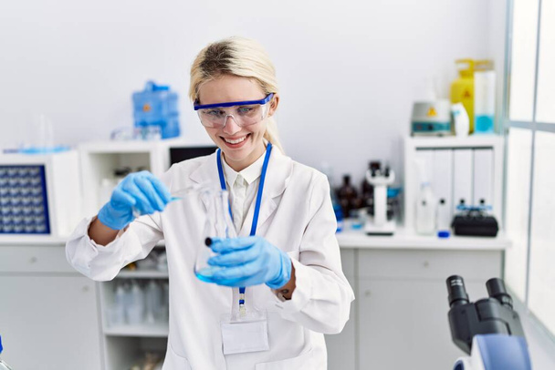 Νεαρή ξανθιά γυναίκα επιστήμονας ρίχνει υγρό στο δοκιμαστικό σωλήνα στο εργαστήριο - Φωτογραφία, εικόνα