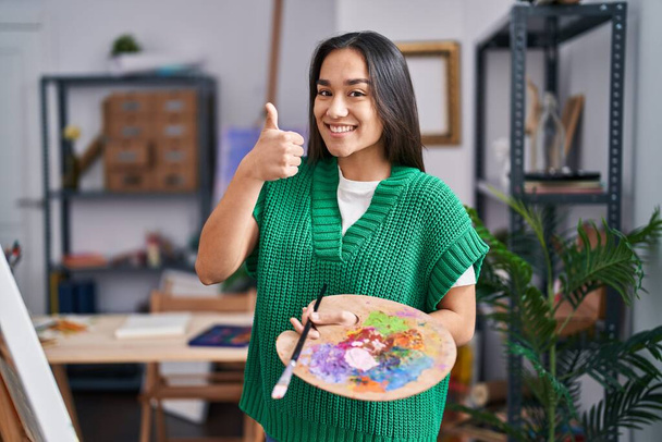 Jovem sul asiático mulher segurando pintor paleta sorrindo feliz e positivo, polegar para cima fazendo excelente e sinal de aprovação  - Foto, Imagem