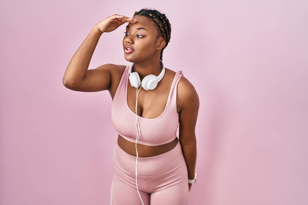 African american woman with braids wearing sportswear and headphones very happy and smiling looking far away with hand over head. searching concept.  - Zdjęcie, obraz