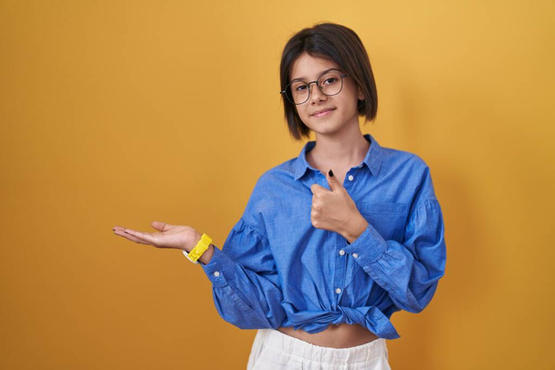 Young girl standing over yellow background showing palm hand and doing ok gesture with thumbs up, smiling happy and cheerful  - Fotó, kép