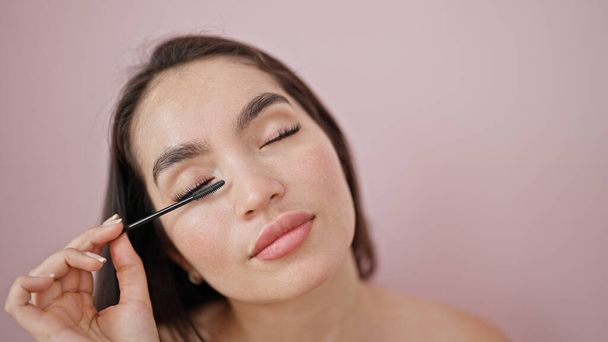 Jonge mooie latino vrouw aanbrengen make-up op wimpers over geïsoleerde roze achtergrond - Foto, afbeelding