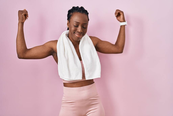 Красивая черная женщина в спортивной одежде и полотенце на розовом фоне показывает мышцы рук, улыбающиеся с гордостью. фитнес концепция.  - Фото, изображение