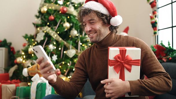 Joven hombre hispano usando smartphone sentado en sofá sosteniendo regalo de Navidad en casa - Foto, imagen