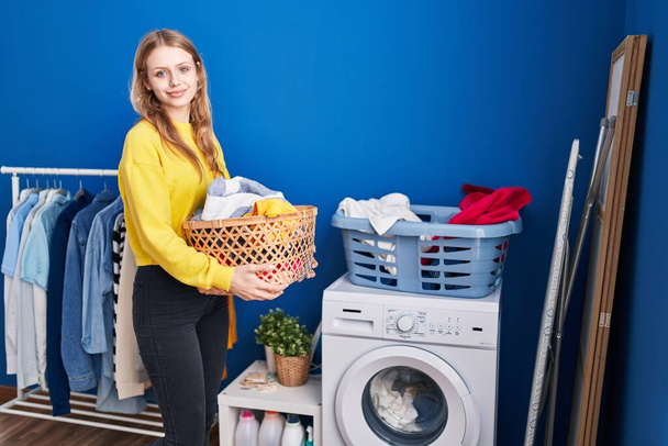 Giovane donna bionda sorridente fiducioso cesto con i vestiti in lavanderia - Foto, immagini