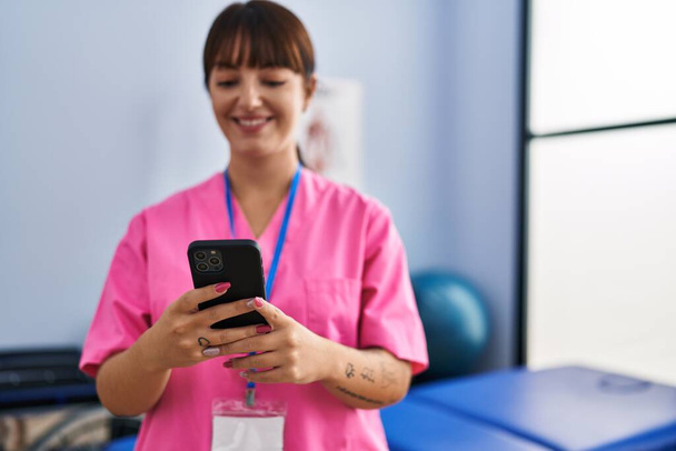 Молодая красивая латиноамериканка физиотерапевт улыбается уверенно, используя смартфон в реабилитационной клинике - Фото, изображение