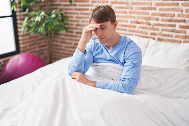 Jonge blanke man gestrest zittend op bed in de slaapkamer - Foto, afbeelding
