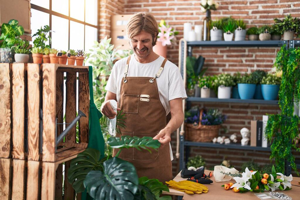 Young blond man florist watering plant working at flower shop - Foto, afbeelding