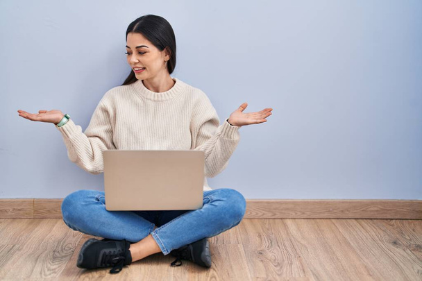 Giovane donna che utilizza il computer portatile seduto sul pavimento a casa sorridente mostrando entrambe le mani palme aperte, presentando e pubblicizzando confronto ed equilibrio  - Foto, immagini