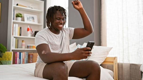 Uomo afroamericano utilizzando smartphone seduto sul letto che celebra in camera da letto - Foto, immagini