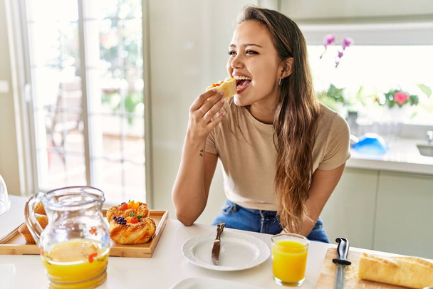 Young beautiful hispanic woman eating bread with jam having breakfast at the kitchen - Photo, Image