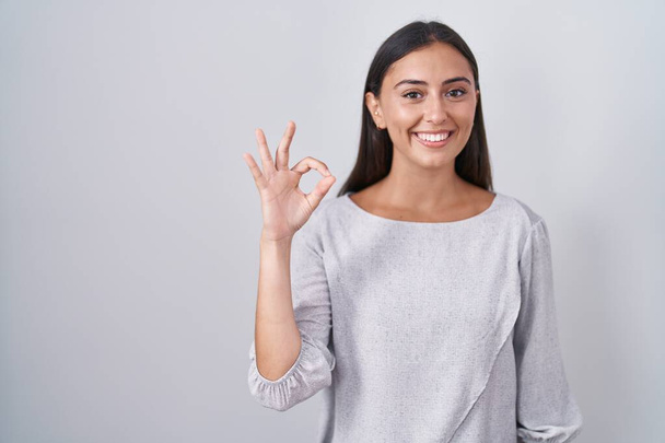 Junge hispanische Frau, die vor weißem Hintergrund steht und positiv lächelt und mit Händen und Fingern Okay-Zeichen macht. Erfolgreicher Ausdruck.  - Foto, Bild