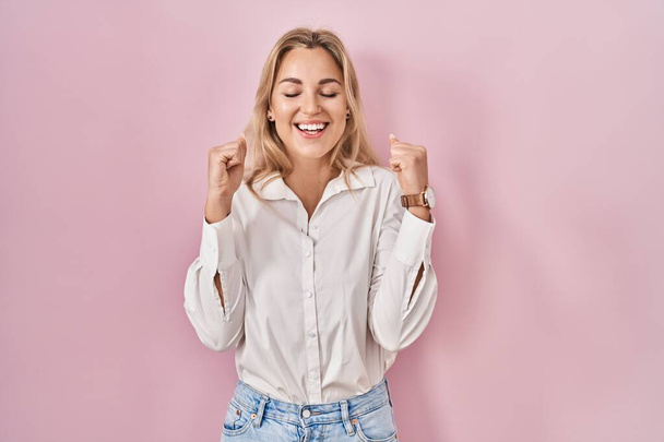 Fiatal kaukázusi nő viselése alkalmi fehér ing felett rózsaszín háttér izgatott a siker karját, és csukott szemmel ünnepli győzelem mosolyogva. győztes koncepció.  - Fotó, kép