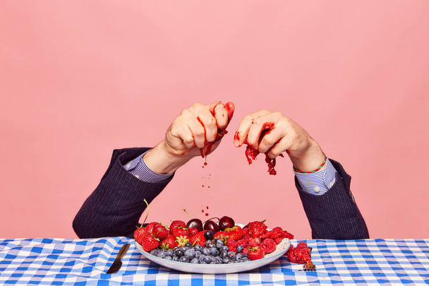 Male hands in classical clothes squeezing fresh berries over plate against pink background. Vitamin smoothie. Concept of food, creativity, party, summer, health. Pop art photography. Copy space for ad - Fotó, kép