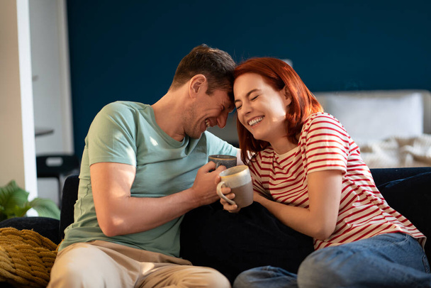 Ευτυχισμένο ξέγνοιαστο μεσήλικο ζευγάρι ερωτευμένο να αστειεύεται γελώντας χαλαρώνοντας στον καναπέ με καφέ, δένοντας, απολαμβάνοντας την πρωινή ώρα μαζί στο σπίτι. Συναισθηματική σύνδεση σε μια σχέση. Έννοια της λαμπρότητας - Φωτογραφία, εικόνα