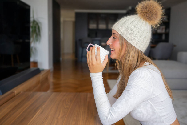 Фото досить мрійливої молодої жінки, одягненої в зимовий капелюх, що сидить на кушетці покриває ковдру, п'є каву в приміщенні домашньої кімнати - Фото, зображення