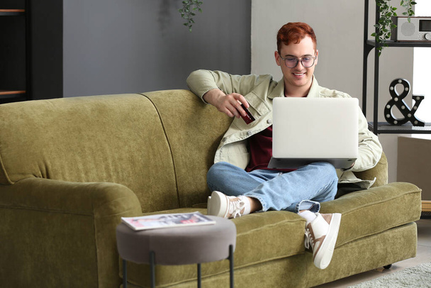 Beau jeune homme avec carte de crédit et ordinateur portable moderne assis sur le canapé tout en faisant des achats en ligne - Photo, image