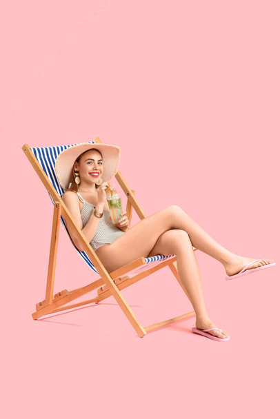 Beautiful young woman in wicker hat with glass of tasty mojito sitting on deckchair against pink background - Photo, Image