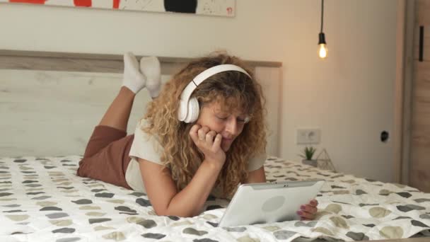Jedna kaukaska kobieta z kręconymi włosami siedzieć w domu za pomocą tabletu cyfrowego i słuchawek do oglądania filmów lub seriali online strumienia lub mieć połączenie wideo szczęśliwy uśmiech mieć dobry czas przestrzeń kopiowania - Materiał filmowy, wideo