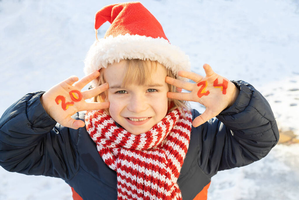 Noel Baba şapkalı ve çizgili kırmızı eşarplı gülümseyen neşeli çocuk, 2024 yılının sayısı avuç içlerine yazılmıştır. Çocuk Noel arifesinde mucize bekliyor. Yeni yıl kış tatili, güneşli soğuk bir gün. - Fotoğraf, Görsel