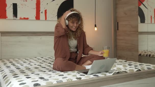 Kıvırcık saçlı beyaz bir kadın evde oturup dijital tablet ve kulaklık kullanarak film ya da dizi izliyor ya da mutlu bir gülümseme videosu çekiyor. - Video, Çekim