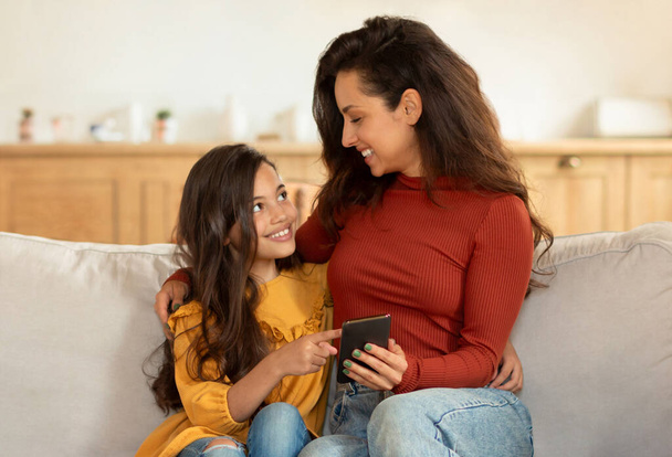 Happy Middle Eastern Mom And Little Daughter Using Smartphone, Kid Girl Pointing Finger At Gadget, Showing Her Favorite Mobile Game Application, Hugging Sitting On Couch At Home Interior - Photo, Image
