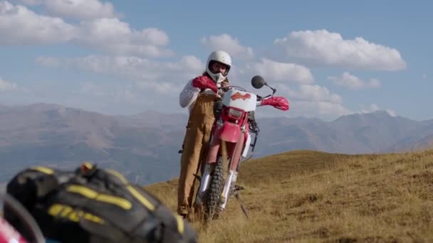 Um motociclista macho remove seu capacete perto de uma motocicleta de enduro off-road vintage, contra o cenário deslumbrante de montanhas e um céu adornado com nuvens. Experimente a emoção de montar um - Filmagem, Vídeo