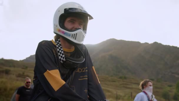 Temidden van een prachtig berglandschap, een jonge man zit op een vintage off-road motorfiets, het aantrekken van een witte helm die zijn avontuurlijke geest onthult. Met de open helm kan de wind - Video