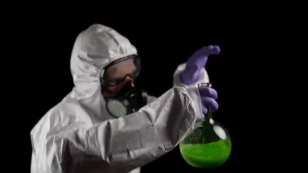 Ein Mann tanzt im Chemikalienschutzanzug mit Atemschutzmaske und Schutzbrille mit grüner Lösung in einem Kolben auf schwarzem Hintergrund - Filmmaterial, Video