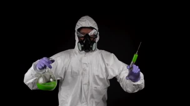 Kimyasal koruma giysisiyle dans eden bir adam solunum cihazıyla ve siyah arka planda şırınga ve matarayla gözlük takıyor. - Video, Çekim