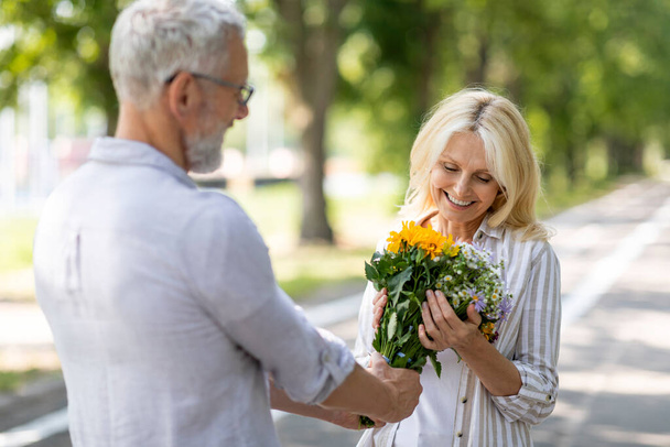Любить зрелого мужчину, дарящего цветы своей жене во время романтического свидания на свежем воздухе, заботиться о старшем муже, удивлять счастливую женщину с букетом, поздравлять с годовщиной или днем рождения, пока они прогуливаются в парке - Фото, изображение