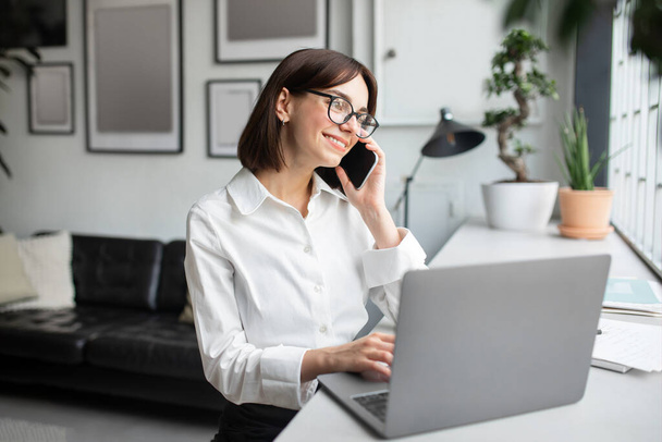 Entspannte Geschäftsfrau, die am Schreibtisch im Büro mit dem Handy spricht, lächelnde Frau, die angenehme Handygespräche führt, während sie am Laptop arbeitet, Kopierraum - Foto, Bild