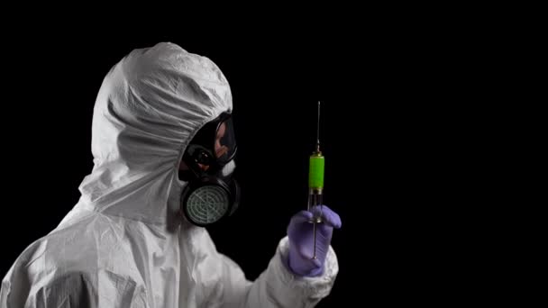 Laboratoriummedewerker in een chemisch beschermingspak houdt een spuit met een groene injectie in zijn handen op een zwarte achtergrond - Video