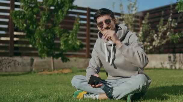 Un tipo guapo con gafas de sol con un teléfono en las manos, se sienta en el césped en el patio, sonríe y mira a la cámara. Comunicación a través de las redes sociales. Citas sitios web. Imágenes de alta calidad 4k - Metraje, vídeo