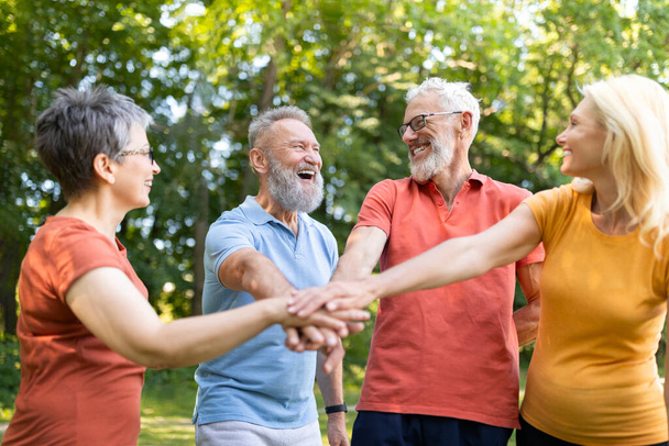 Motywacja grupowa. Szczęśliwi seniorzy w odzieży sportowej układanie rąk po treningu na świeżym powietrzu, radośni dojrzali mężczyźni i kobiety witają się z sukcesem w pracy zespołowej, ciesząc się aktywnym stylem życia - Zdjęcie, obraz