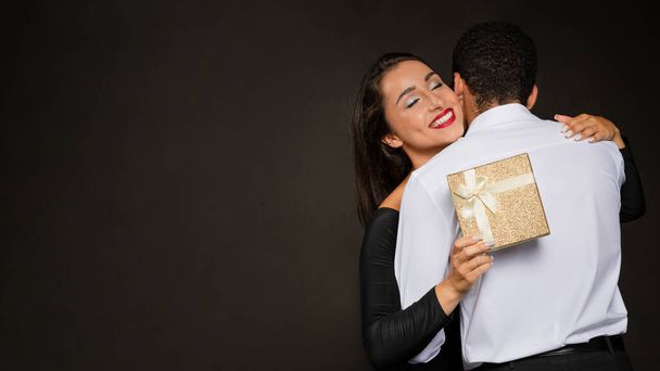 Happy beautiful brunette young woman in nice black dress and bright makeup hold gift box present and hugging boyfriend, couple celebrating xmas, new year, anniversary, birthday on black, copy space - Photo, Image