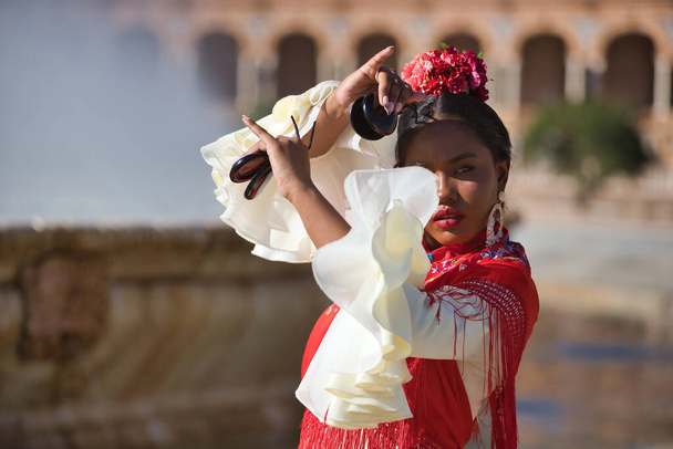 Young black and South American woman in a beige gypsy flamenco suit and red shawl, dancing with castanets in a beautiful square in the city of Seville in Spain. Concept dance, folklore, flamenco, art - Photo, Image