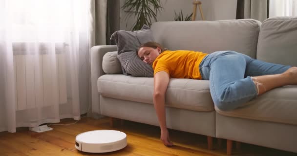 Πολύ κουρασμένη γυναίκα κοιμάται στον καναπέ στο σπίτι, ενώ η ηλεκτρική σκούπα ρομπότ εργάζεται στο πάτωμα. Υψηλής ποιότητας 4k πλάνα - Πλάνα, βίντεο