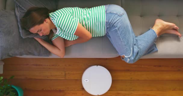 Mulher muito cansada dormindo no sofá em casa, enquanto robô aspirador de pó trabalhando no chão. Imagens 4k de alta qualidade - Filmagem, Vídeo