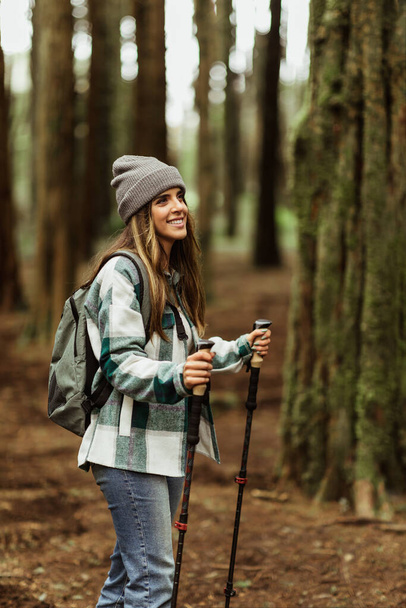 Glad young caucasian lady tourist in jacket with trekking sticks walk in forest alone, enjoy trip, outdoor, vertical. Travel, hiking, active lifestyle, adventure and tourism - Photo, Image